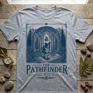 May include: A heather gray t-shirt featuring a blue graphic of a dog running through a forest. The text "THE PATHFINDER" is below the graphic, with "LOYAL. WISE. WILD." underneath. The shirt has a crew neck and short sleeves.