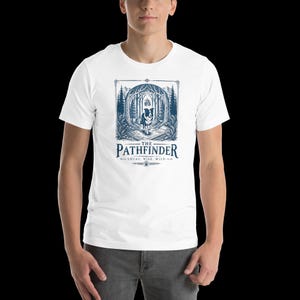 May include: White t-shirt featuring a detailed blue illustration of a dog walking through a forest, with the text "THE PATHFINDER" and the words "LOYAL. WISE. WILD." below the image.