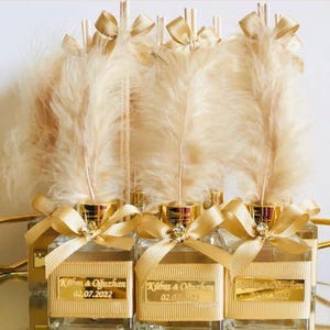 May include: Clear glass bottles with gold accents, ribbons, and feathers. Each bottle has a gold label with the text "Kibum & Ouzhan" and the date "02.07.2022". The bottles are arranged in a row, suggesting a wedding or event favor.