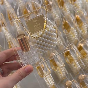 Pode incluir: Caixas de lembrancinhas de acrílico transparente com detalhes dourados e enfeites de pérolas. Cada caixa contém um colar de pérolas, uma pequena garrafa dourada e um objeto decorativo transparente. As caixas estão dispostas em uma bandeja, com o texto "Sibel & Celal" em um rótulo dourado.