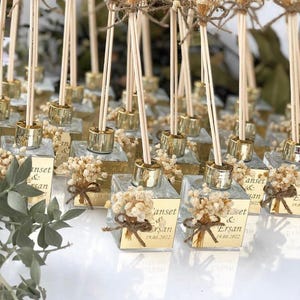Pode incluir: Uma coleção de pequenos frascos de vidro transparente com tampas douradas e varetas de madeira. Cada frasco é adornado com um pequeno buquê de flores secas e uma etiqueta dourada com o texto "Anset & Ersan 19.08.2022".