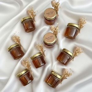 May include: Small glass jars filled with honey, each topped with a gold lid and a wooden honey dipper. The jars are decorated with dried flowers and a small label. The jars are arranged on a white satin surface.