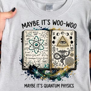 Pode incluir: Camiseta cinza claro com as frases "MAYBE IT'S WOO-WOO" e "MAYBE IT'S QUANTUM PHYSICS". O design apresenta um livro aberto com diagramas científicos e símbolos místicos, como um átomo e um olho que tudo vê.