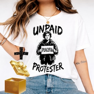 Może przedstawiać: Biała koszulka z czarno-białą grafiką przedstawiającą osobę trzymającą znak z napisem „PEACEFUL”. Na koszulce widnieją również słowa „UNPAID PROTESTER”. Złote pudełko na prezent i czarny krzyż również znajdują się na zdjęciu.
