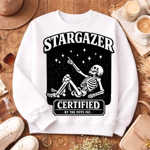 Op de afbeelding: Witte sweatshirt met een zwarte afbeelding van een skelet dat naar sterren wijst. De tekst "STARGAZER" staat boven de afbeelding en "CERTIFIED" en "BY THE POTS INC." eronder. De sweatshirt ligt op een houten oppervlak.
