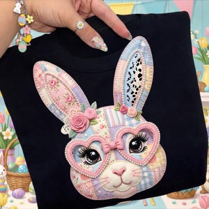 May include: A black sweatshirt featuring a colorful patchwork bunny face with heart-shaped glasses and a pink bow. The bunny has large eyes, long eyelashes, and is decorated with pink roses and lace details. The design is centered on the front of the shirt.