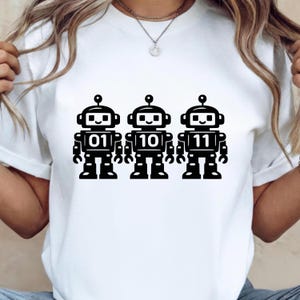 May include: White t-shirt featuring three black robot designs. Each robot has a different number on its chest: 01, 10, and 11. The robots have antenna, square heads, and smiling faces. The shirt is made of a soft material.