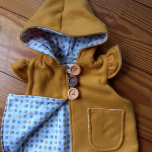 May include: A mustard yellow hooded vest with a white and blue polka dot lining. The vest has a pocket on the front and three buttons.