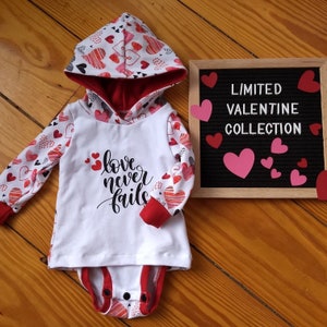 May include: White hooded onesie with red trim and a heart pattern. The onesie has the words "love never fails" printed on the front. The onesie is part of a limited Valentine's Day collection.