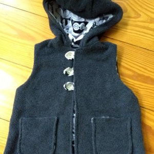 May include: A gray hooded vest with two pockets and button closures. The vest is lined with a black and white patterned fabric.