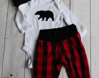 May include: A baby outfit set featuring a red and black checked pattern. The set includes a long-sleeved bodysuit with a black bear graphic, matching trousers, and a beanie hat.