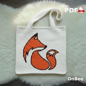 Puede incluir: Bolso tote blanco con un diseño de zorro tejido en naranja y negro. El bolso tiene forma rectangular con un asa y el texto "PDF" y "OnBee". El bolso está sobre una superficie blanca y esponjosa.