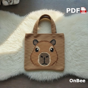 Könnte beinhalten: Eine hellbraune Häkeltasche mit einem Capybara-Gesicht. Das Capybara hat braune und schwarze Details. Die Tasche hat zwei Henkel und der Text "OnBee" ist sichtbar. Die Tasche liegt auf einem weißen, flauschigen Teppich.