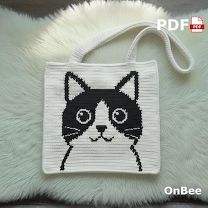 May include: A white crocheted tote bag featuring a black and white cat face design. The bag has a rectangular shape with a handle and the text "PDF" and "OnBee". The cat's face is centered on the front of the bag.