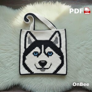 Puede incluir: Bolso tote tejido a ganchillo en blanco y negro con la cara sonriente de un perro husky con ojos azules. El bolso tiene un asa blanca y un borde negro. El texto "PDF" y "OnBee" son visibles.