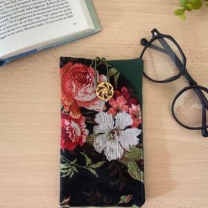 Può includere: Un astuccio per occhiali in tessuto arazzo floreale con sfondo nero, con fiori rossi, bianchi e verdi. L'astuccio ha una tasca verde ed è assicurato con una chiusura color oro. Un paio di occhiali con montatura nera è nelle vicinanze.