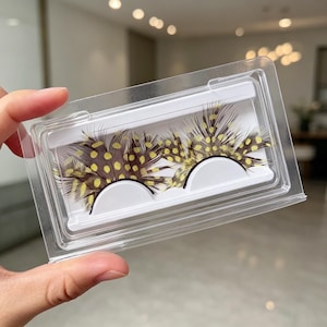 May include: A pair of false eyelashes made from brown feathers with yellow polka dots, presented in a clear rectangular plastic storage case with a white interior tray that holds each lash in a curved position for display and protection.