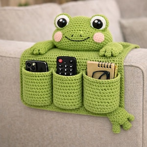 May include: A green crocheted frog-shaped couch organizer with three pockets. The frog has large black and white eyes, pink cheeks, and outstretched arms. The pockets hold a phone, remote control, notebook, and glasses. The organizer is designed to drape over a couch arm.