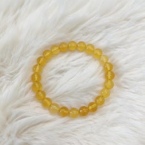 Op de afbeelding: Een cirkelvormige elastische armband bestaande uit gladde, ronde, doorschijnende gele kralen. De kralen vertonen een consistente goudgele tint met subtiele variaties in helderheid en lichtreflectie over de gepolijste, bolvormige oppervlakken.