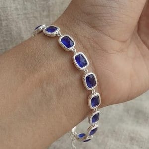 May include: A silver-toned bracelet featuring a series of rectangular links, each set with a deep blue stone. The bracelet has a textured metal finish and includes an adjustable chain extension with a small circular charm at the end.