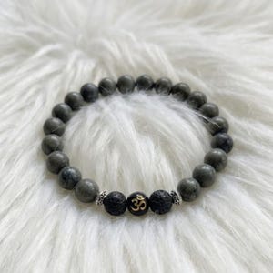 May include: A beaded bracelet featuring smooth, round grey stone beads, two textured black lava beads, and a central black bead with a gold-colored Om symbol. The bracelet includes small silver-toned metal spacers between the focal beads.