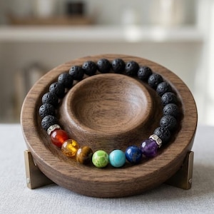 May include: A beaded bracelet featuring black porous lava stone beads and seven smooth, colorful chakra stones including red, orange, brown, green, light blue, dark blue, and purple, accented by two silver-toned metal spacers with small clear crystals.