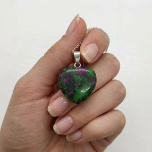 May include: A heart-shaped pendant necklace with a polished gemstone. The pendant, a mix of green and purple, hangs from a delicate silver chain. The gemstone is held by a silver bail. The necklace is a piece of jewellery.