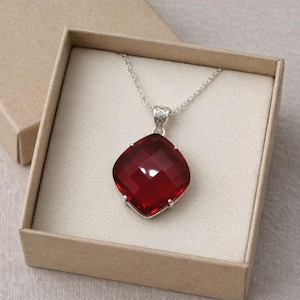 May include: A ruby-red, faceted gemstone pendant necklace with a silver chain, displayed in a beige gift box. The pendant is a large, square shape with a brilliant cut. The open box reveals the necklace, ready for gifting.