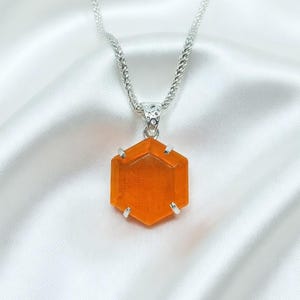 May include: A silver necklace featuring a hexagonal orange pendant. The pendant, a vivid orange hue, hangs from a silver chain. The necklace is presented against a white satin backdrop.