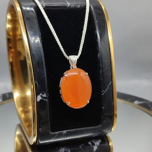 May include: A silver necklace featuring an oval, orange gemstone pendant. The pendant is set in a silver frame and hangs from a delicate silver chain. The necklace is displayed on a black and gold jewelry box.