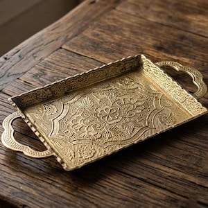 May include: A rectangular brass-colored metal tray featuring ornate embossed floral patterns across the base and edges. The tray includes two decorative handles on opposite sides, with a textured finish throughout the entire surface area.