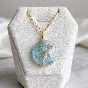 Op de afbeelding: Een halvemaanvormige hanger van lichtblauwe doorschijnende steen, omwikkeld met goudkleurig draad in de vorm van een boom. De hanger hangt aan een fijne goudkleurige ketting en is voorzien van gedetailleerde draadvertakkingen over de steen.