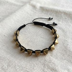 May include: A black cord bracelet featuring a series of metallic gold-toned pyramid-shaped studs arranged along the band. The bracelet includes an adjustable closure mechanism with two small dark beads at the ends of the black cord strings.