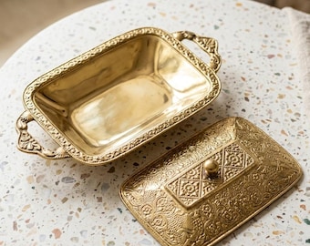 May include: A rectangular brass butter dish with ornate handles and a matching lid. The lid features an intricate embossed geometric and floral pattern with a central knob, while the base has a smooth interior and decorative edges.
