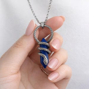 May include: A silver necklace with a dark blue crystal pendant. A detailed snake design wraps around the crystal. The pendant hangs from a silver chain. The crystal is a deep blue colour.