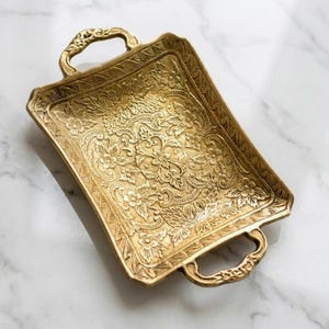 May include: A rectangular brass tray featuring an intricate embossed floral pattern across its surface. The tray includes two decorative handles on opposite ends, with a textured, metallic finish that highlights the detailed botanical engravings.