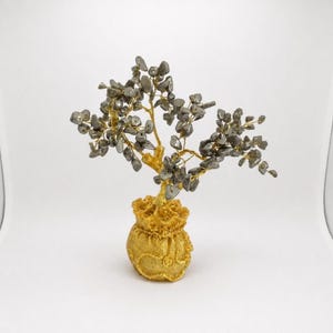 May include: A decorative tree sculpture featuring numerous small, tumbled grey stones attached to thin, twisted gold-colored wire branches. The tree is anchored in a textured, gold-colored base shaped like a small, rounded pouch or sack.