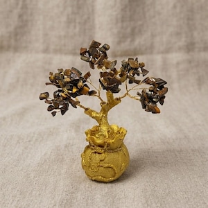 Puede incluir: Una escultura de árbol decorativa con fragmentos de piedra ojo de tigre marrón y dorado pulidos unidos a ramas de alambre fino de color dorado. El árbol está anclado en una base redonda texturizada de color dorado con forma de bolsa.