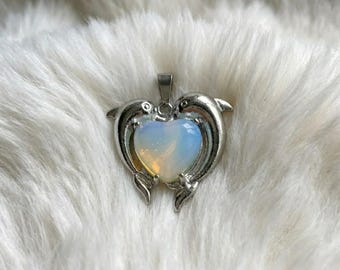 May include: A silver-toned pendant featuring two dolphins embracing a heart-shaped opal. The opal has a gradient of colors, from white to yellow to blue. The pendant is designed to be worn as a necklace.