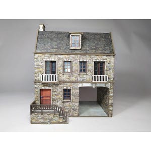 Könnte beinhalten: Ein Miniatur-Steinhaus mit zwei Etagen und einem grauen Schieferdach. Das Haus verfügt über mehrere Fenster, eine rote Tür und einen kleinen Balkon. Eine garagenartige Öffnung ist auf der unteren Ebene sichtbar. Das Haus hat ein rustikales, gealtertes Aussehen.
