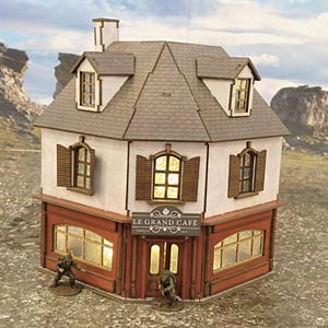 Può includere: Modello in miniatura di un caffè con facciata bianca, persiane marroni e tetto grigio. Il caffè ha una tenda da sole rossa e un'insegna "LE GRAND CAFÉ". Due piccole figure sono davanti al caffè.