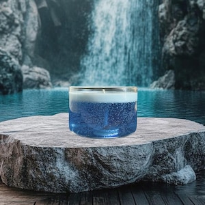 Può includere: Una candela di vetro trasparente con una base di cera blu e frizzante e uno strato superiore bianco. La candela è su una superficie di pietra grigia, con una cascata e un lago sullo sfondo. La candela ha una singola stoppino.