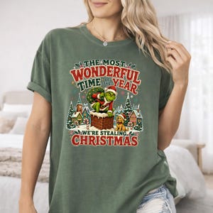 May include: A sage green short-sleeve t-shirt featuring a graphic of a green character in a Santa suit emerging from a chimney with a dog. The text reads: THE MOST WONDERFUL TIME OF THE YEAR WE'RE STEALING CHRISTMAS, surrounded by snowy houses.