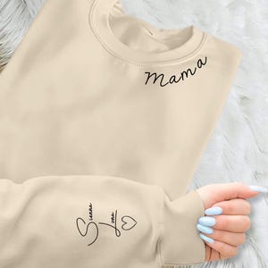 Könnte beinhalten: Ein cremefarbenes Sweatshirt mit dem Wort "Mama" in schwarzer Schrift auf der oberen rechten Brust. Der Ärmel zeigt den Namen "Sienna Luna" in Schrift mit einem Herzsymbol. Das Sweatshirt ist ein lässiges Kleidungsstück für jeden Tag.