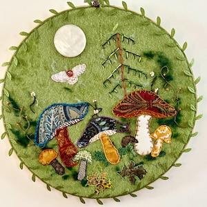 May include: A circular embroidery hoop art piece featuring a green felt background with fabric-appliqued mushrooms in various patterns and colors. A white circular button moon, a small moth, and a stitched tree are present, with leaf trim.
