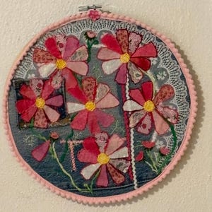 May include: A handmade floral wall hanging. The artwork features pink and red fabric flowers with yellow centers, arranged on a denim background. The design is framed by a pink pom-pom trim and a white lace border. A small pink flower is at the top.