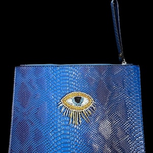 May include: A blue clutch bag with a snakeskin effect and an embroidered eye motif. The eye has a gold border, a blue iris, and gold and white beadwork. A wrist strap is attached to the zip.