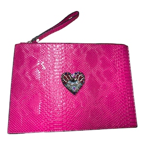 Może przedstawiać: Jasnoróżowa kopertówka o fakturze skóry węża z paskiem na nadgarstek. Kopertówka posiada zdobienie w kształcie serca z kolorowymi kamieniami szlachetnymi i wzorem oka.