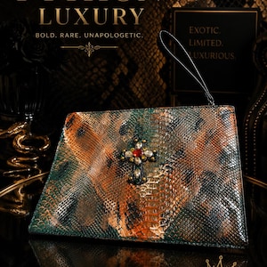 May include: A rectangular clutch bag with a textured, multi-colored snakeskin pattern in shades of green, orange, and black. The front features a central cross embellishment with a red stone and clear crystals. It includes a thin wrist strap.
