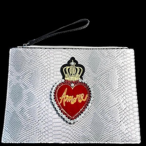 May include: Silver clutch bag with a snakeskin effect, featuring a red heart-shaped emblem with the word "Amore" in gold script. The emblem is adorned with a crown and rhinestones, and has a black wrist strap.